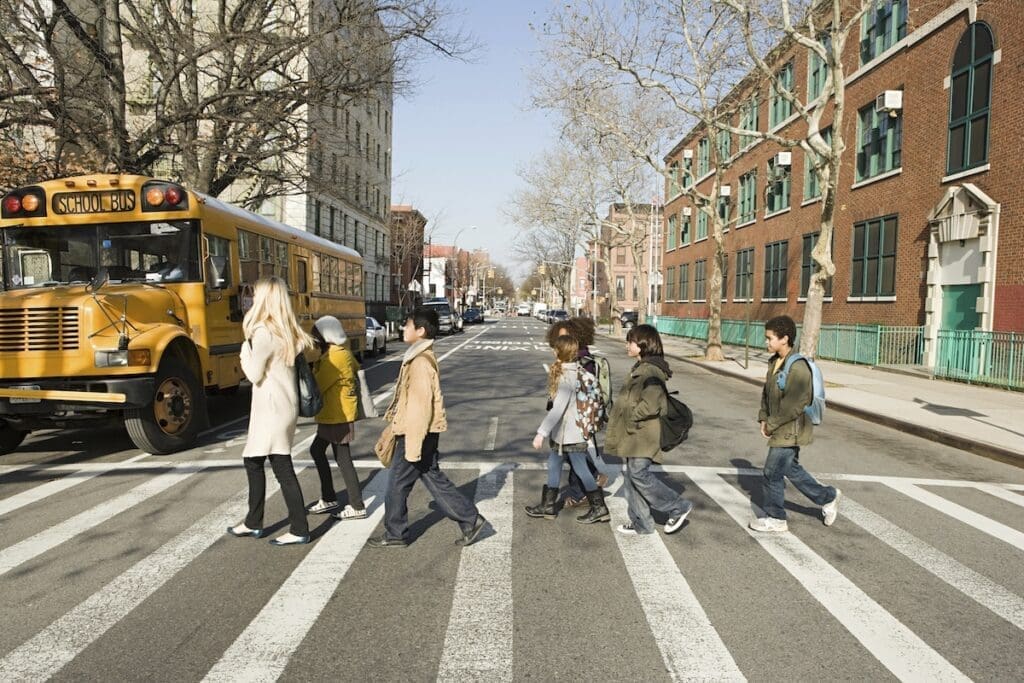 A group of children crossing the street at a pedestrian crosswalk with a yellow school bus waiting nearby and buildings lining the street.